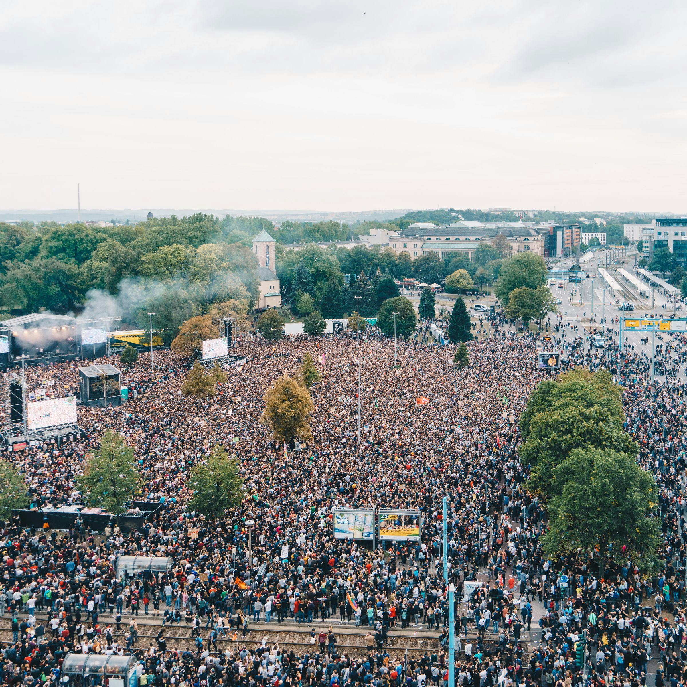 On voit une photo du KOSMOS 2018, au-dessus de laquelle une foule (photographiée d'en haut) remplit entièrement l'espace devant la scène. Les rues adjacentes sont également remplies de gens.
