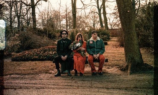 The three band members are sitting on a bench in an autumnal park wearing sunglasses. The singer is holding a bouquet of flowers in her hand.