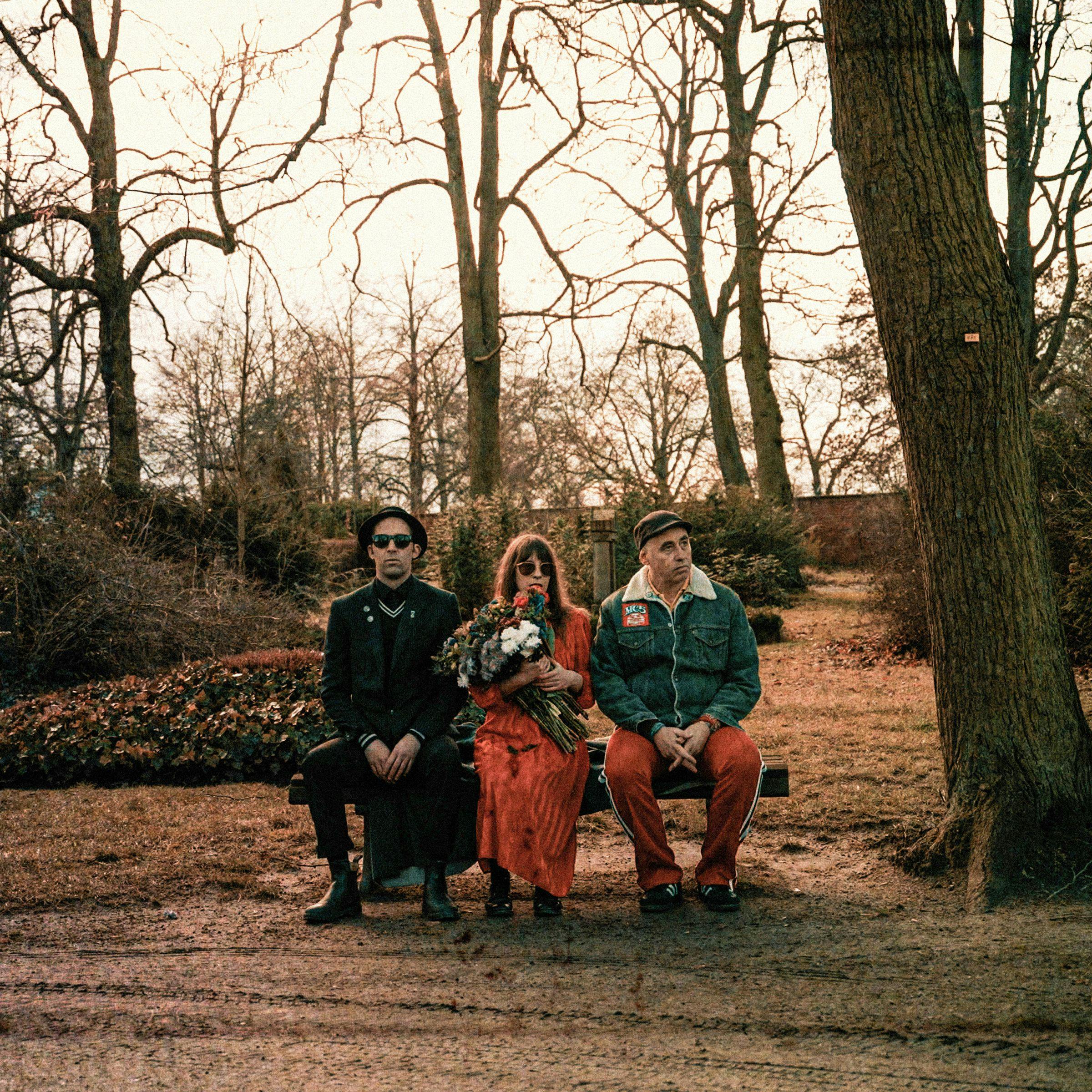 Три участника группы сидят на скамейке в осеннем парке в солнцезащитных очках. Певица держит в руке букет цветов.