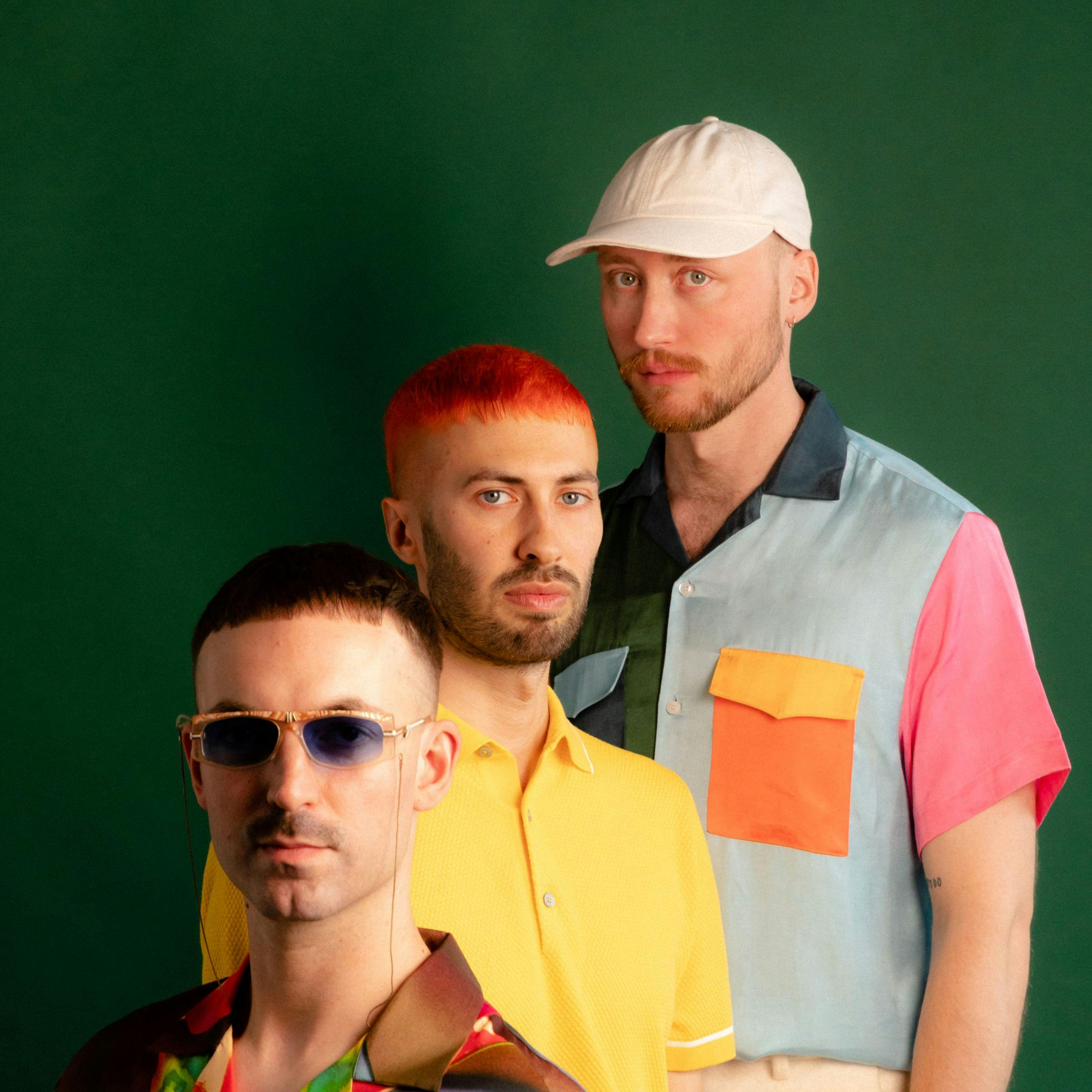 Los tres miembros de la banda Tropikel Ltd. están parados con ropa colorida frente a un fondo verde oscuro mirando directamente a la cámara.