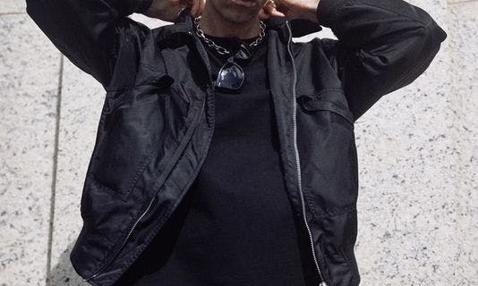Электро-диджей Boys Noize (настоящее имя Алекс Ридха) позирует в черной одежде на фоне бежевой стены, держа руки на воротнике. Он смотрит в камеру.