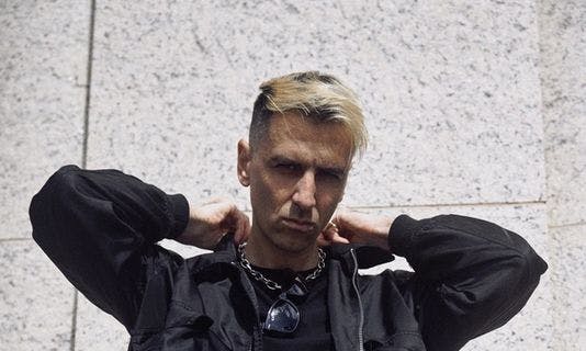 Electro-DJ Boys Noize, whose real name is Alex Ridha, poses dressed in black in front of a beige wall with his hands on his collar. He looks directly into the camera.