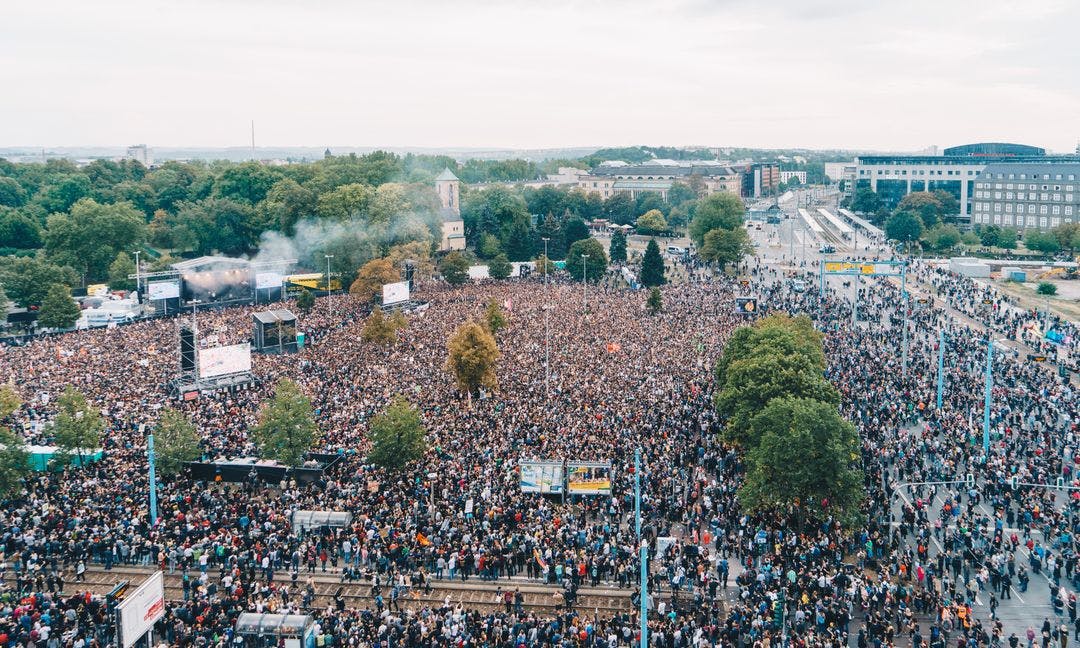 One can see a photo of COSMOS 2018, above which a crowd of people (photographed from above) completely fills the space in front of the stage. The streets adjacent are also filled with people.