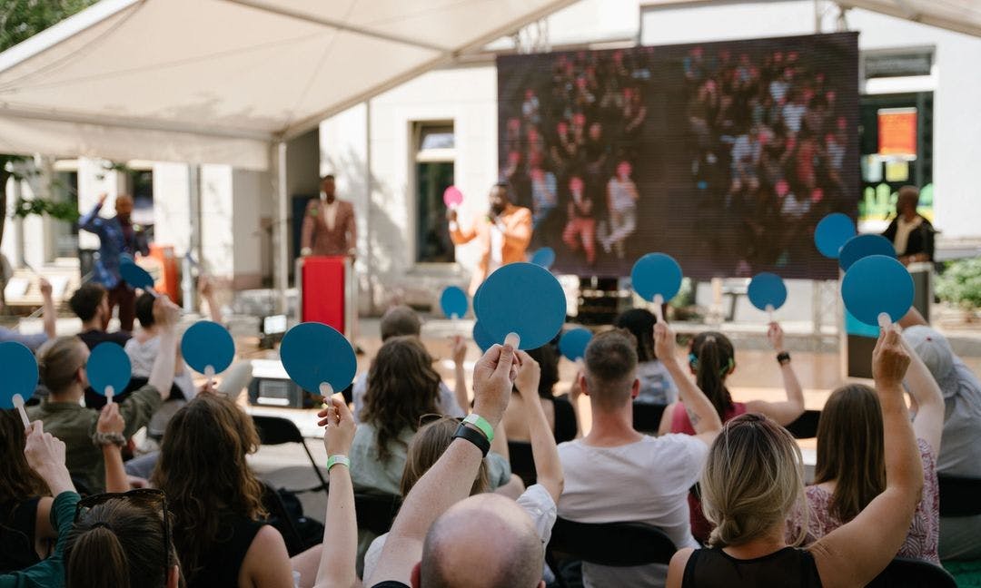 Lors d'un débat en plein air, le public est photographié de dos en train de lever des cartes bleues. À l'arrière-plan, on peut voir la scène avec les animateurs.