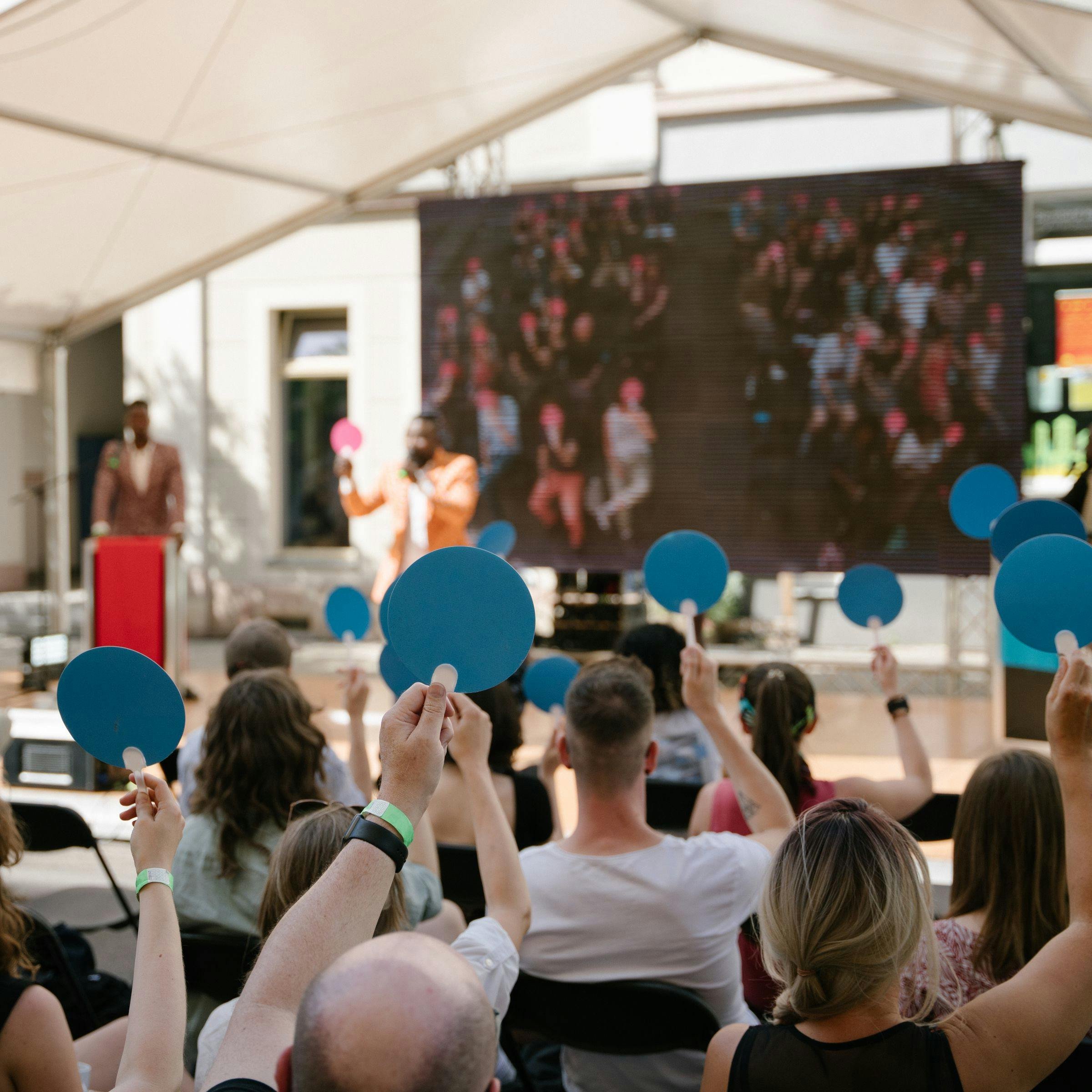 Bei einem Diskussionsformat im Freien wird das Publikum von hinten dabei fotografiert, wie sie blaue Karten nach oben halten. Im Hintergrund sieht man die Bühne mit den Moderatoren.
