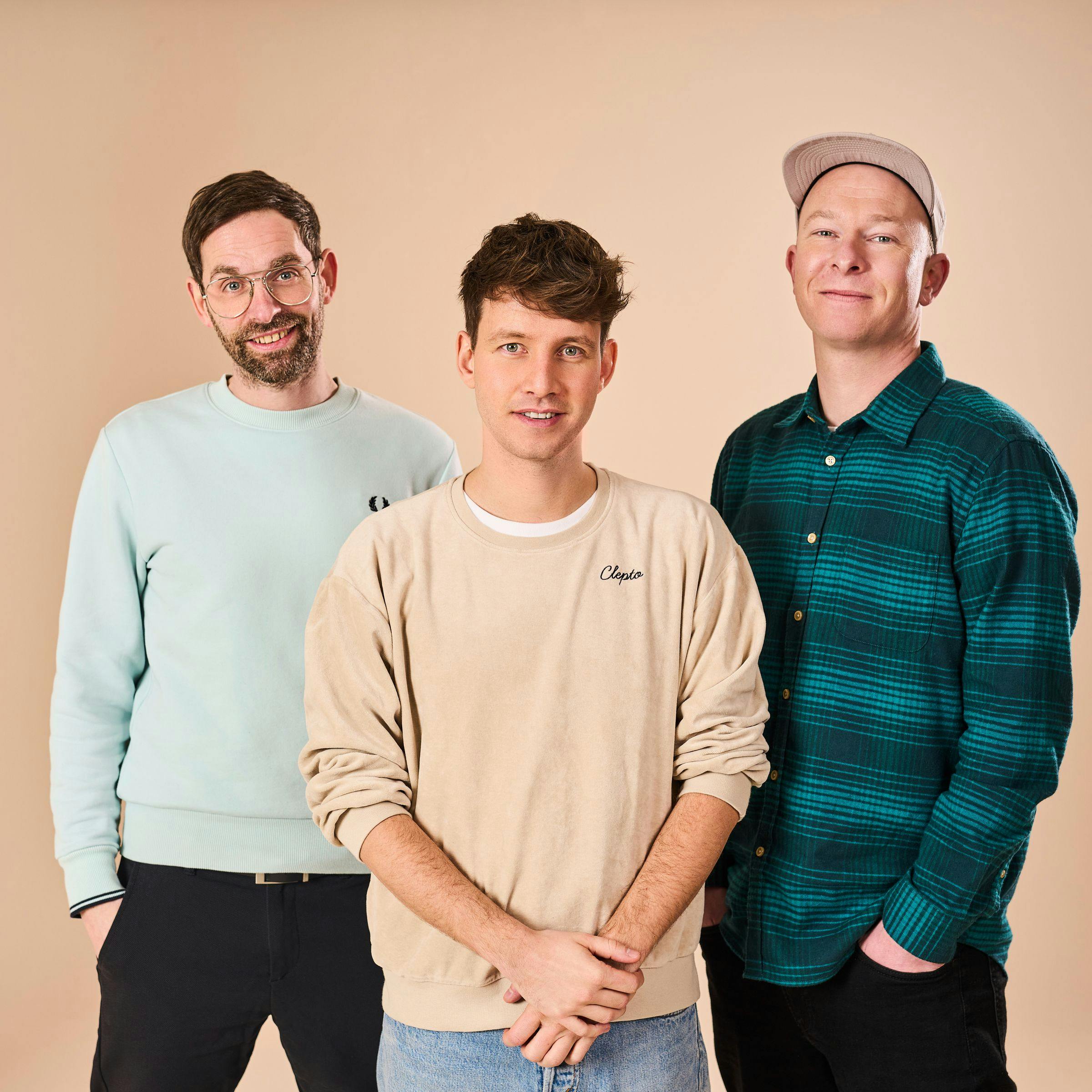 All three members of Deine Freunde are standing side by side in front of a beige-orange wall, looking amiably into the camera.